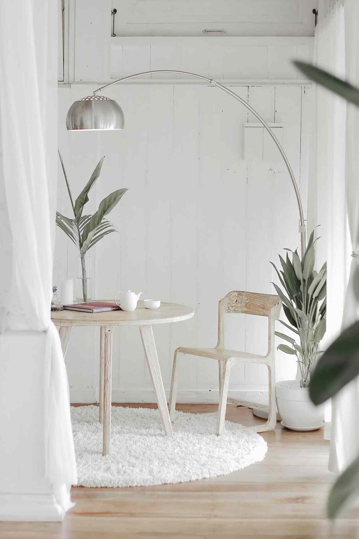 A minimalist home interior featuring a round wooden table, a light-colored chair, a modern arc floor lamp, and a plant beside the table, set against a neutral wall.