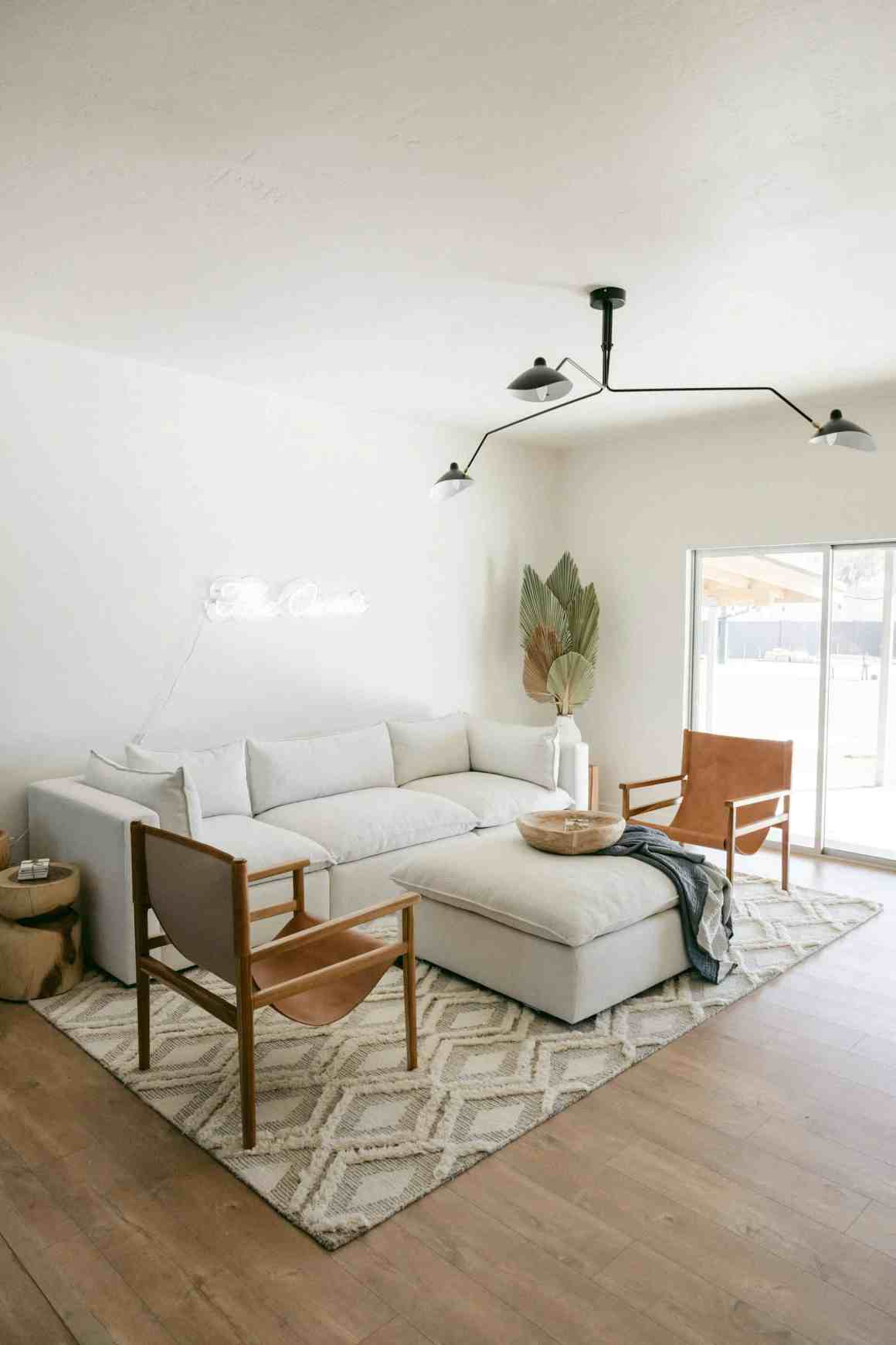 A modern minimalist living room featuring a large white sectional sofa, natural wood accent chairs, a geometric patterned area rug, and a stylish ceiling light fixture.
