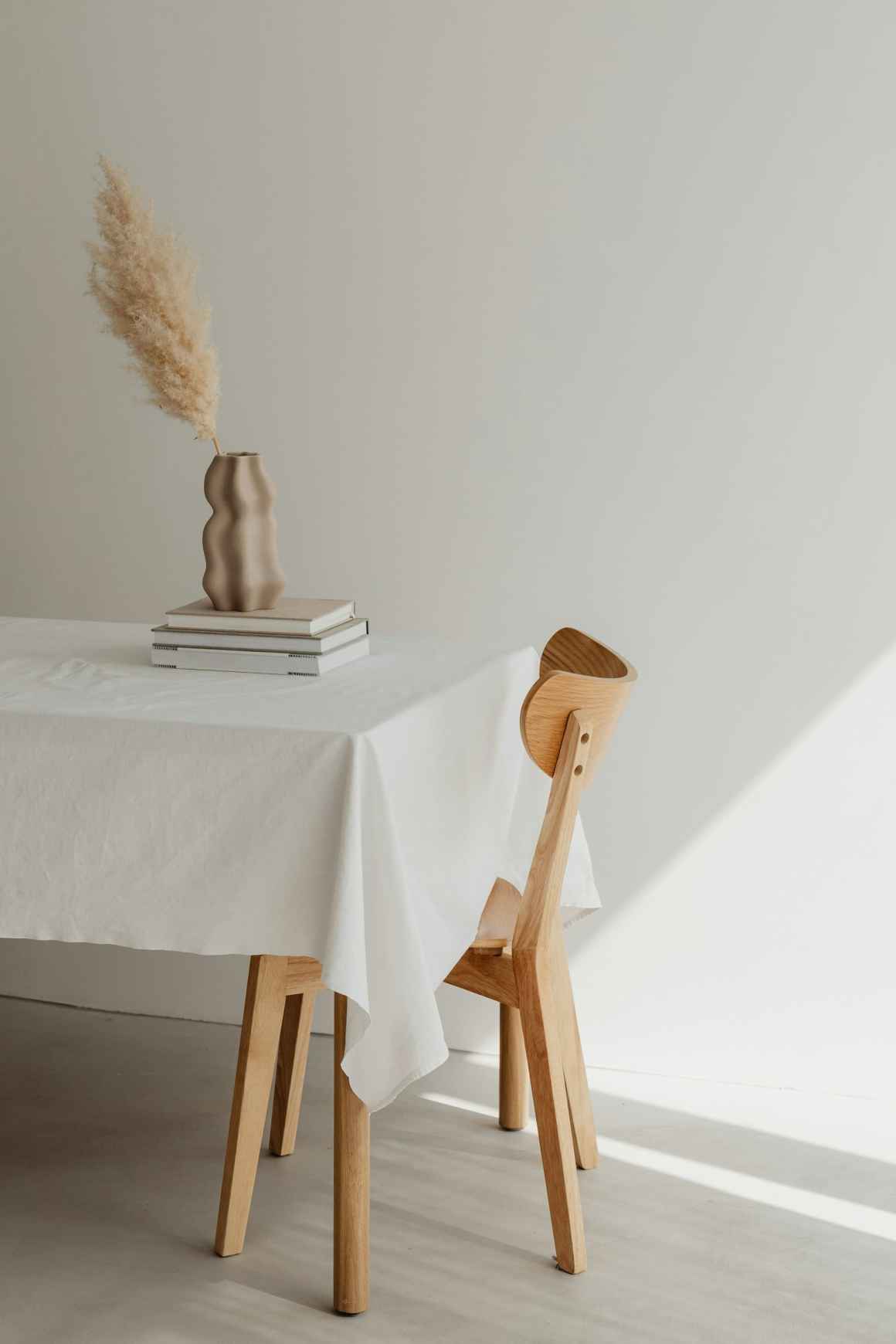 A minimalistic dining setup featuring a wooden chair, a white tablecloth, stacked books, and a decorative vase with dried flowers against a light-colored wall.