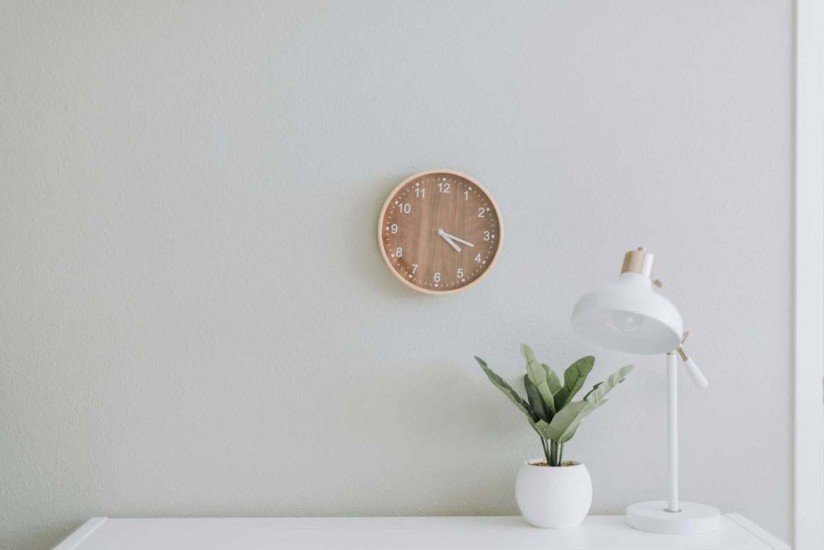 A minimalist interior featuring a wooden wall clock, a small potted plant, and a white desk lamp against a light gray wall.