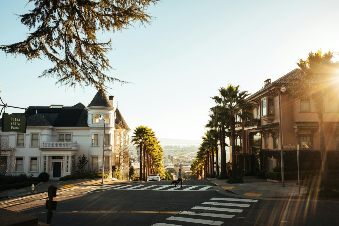 A scenic view of a San Francisco street leading downhill, lined with palm trees and featuring historic architecture, with the sun shining in the background.
