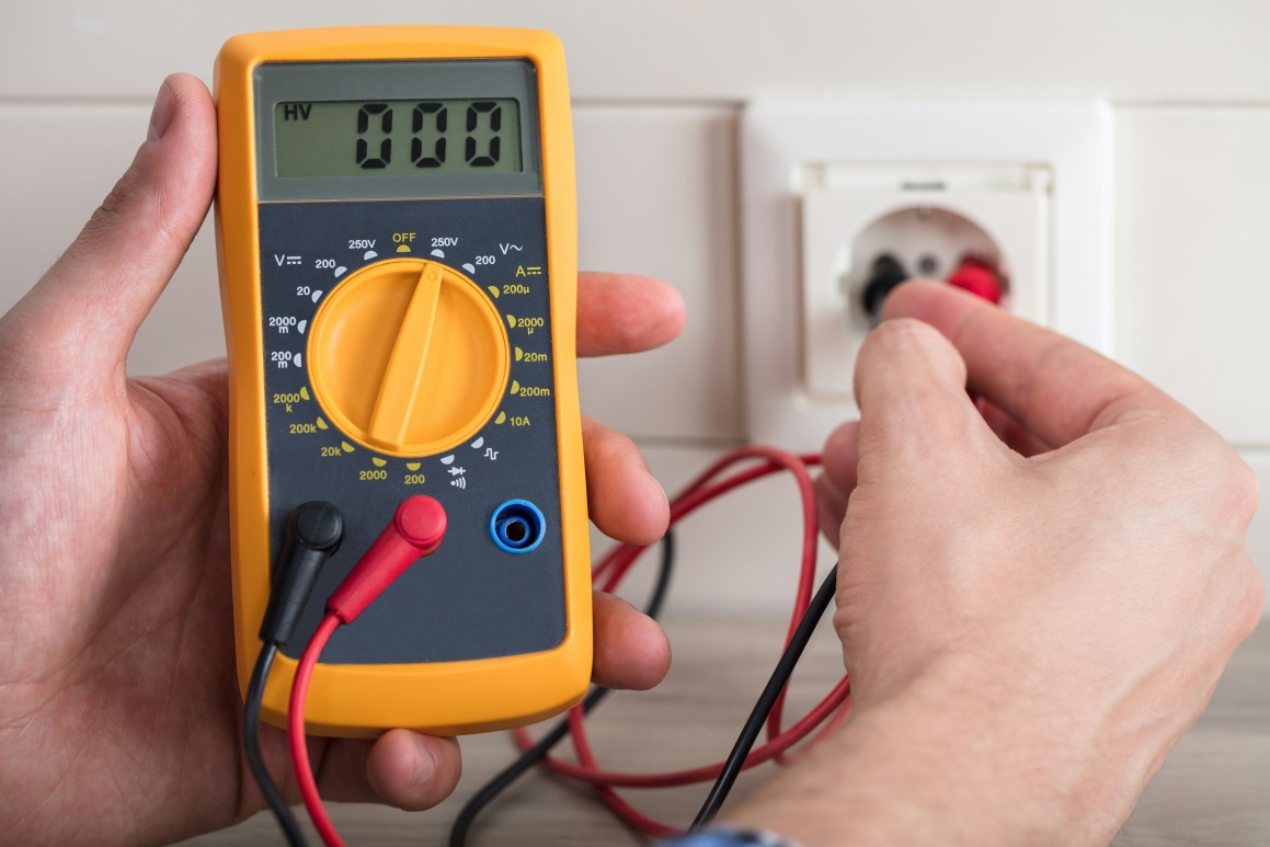 A person using a multimeter to check a wall outlet, with test leads connected to the device and the outlet.