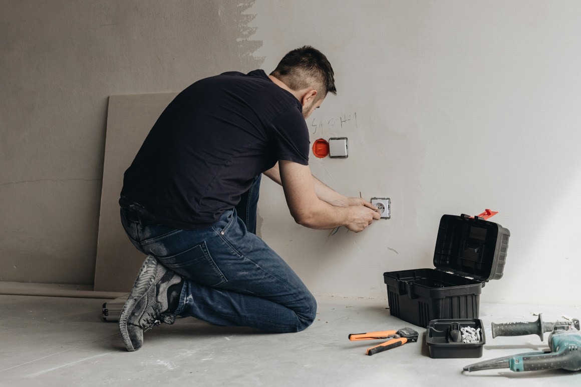 A person kneeling while installing an electrical outlet on a wall, with a toolbox and various tools lying on the floor.