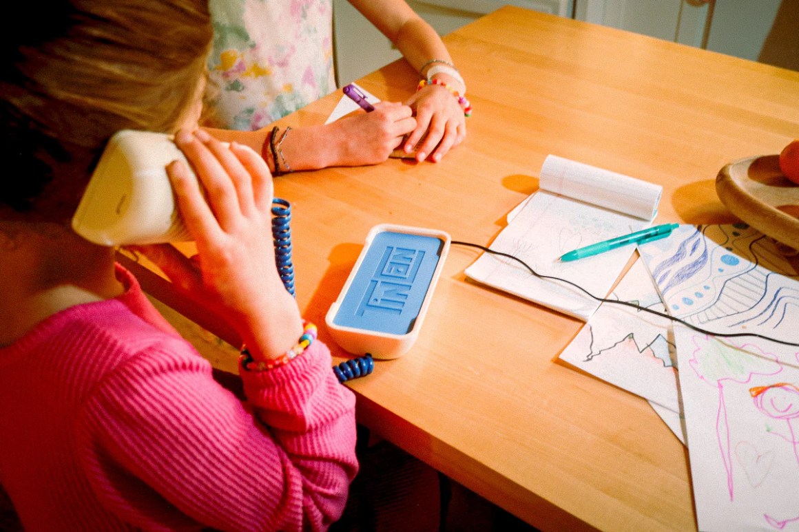 A child holding a retro landline phone while another child is coloring at a table, surrounded by drawings and art supplies.