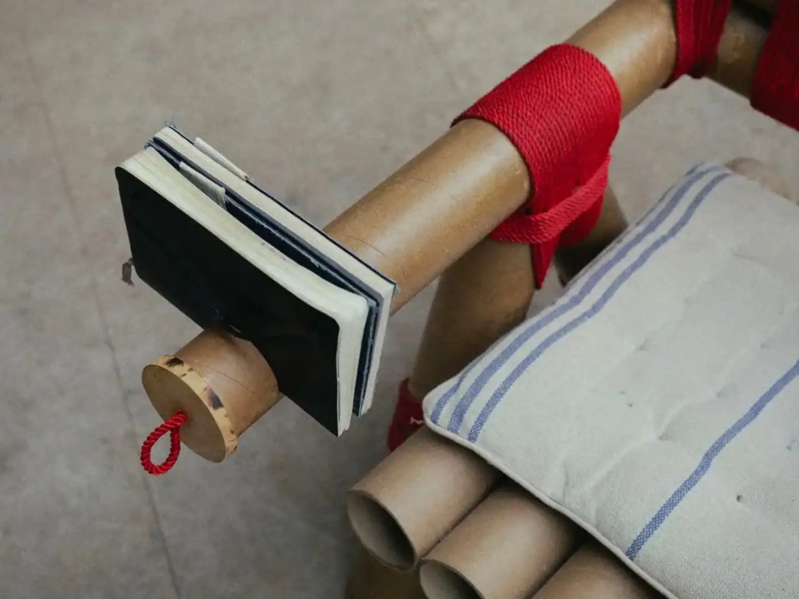 A close-up of a unique chair arm featuring a small notebook attached to a cylindrical support, wrapped in red fabric. The chair has a cushioned seat with horizontal stripes.