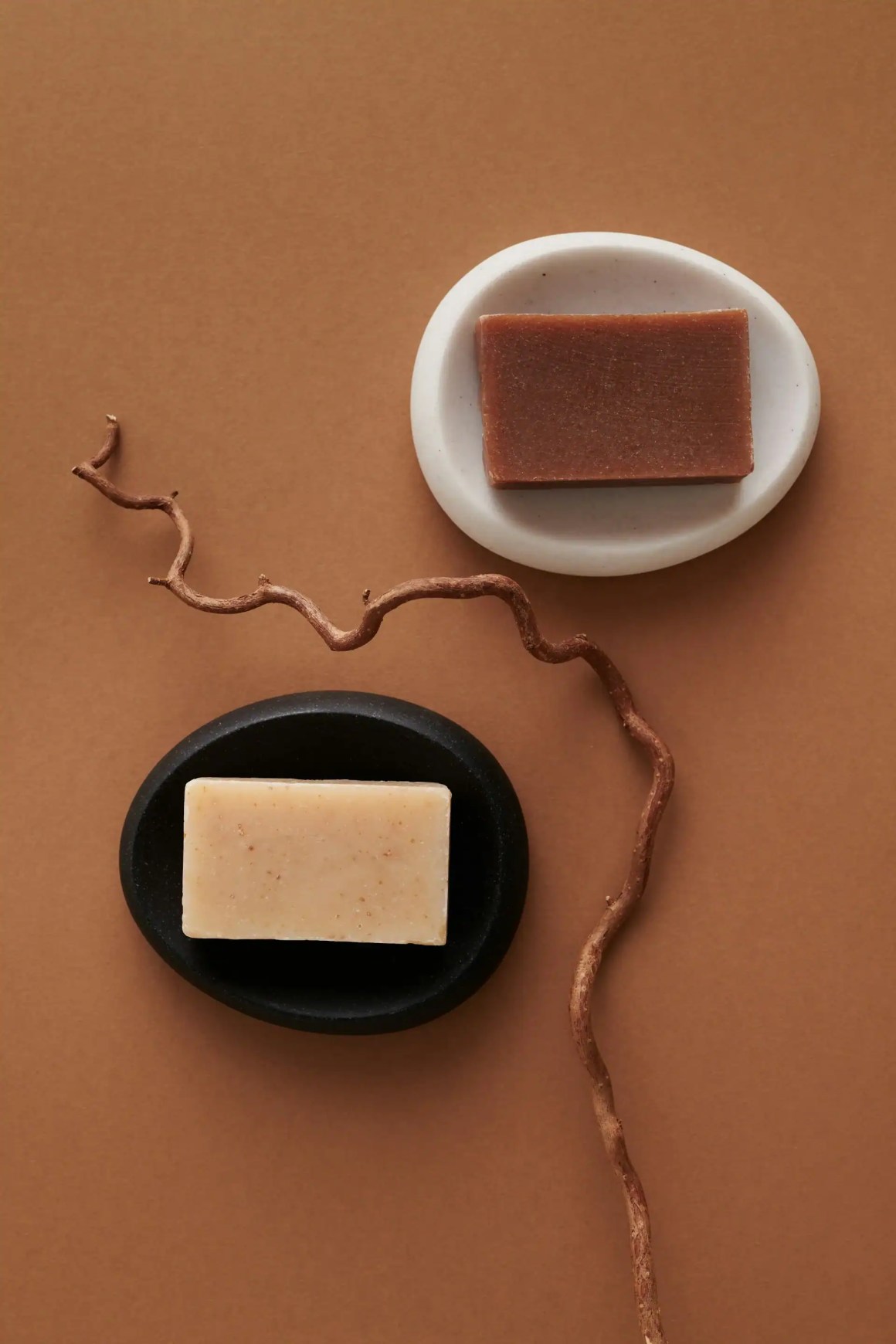 Two bars of soap, one dark brown on a white dish and one light beige on a black dish, placed on a brown background with a twisted branch.