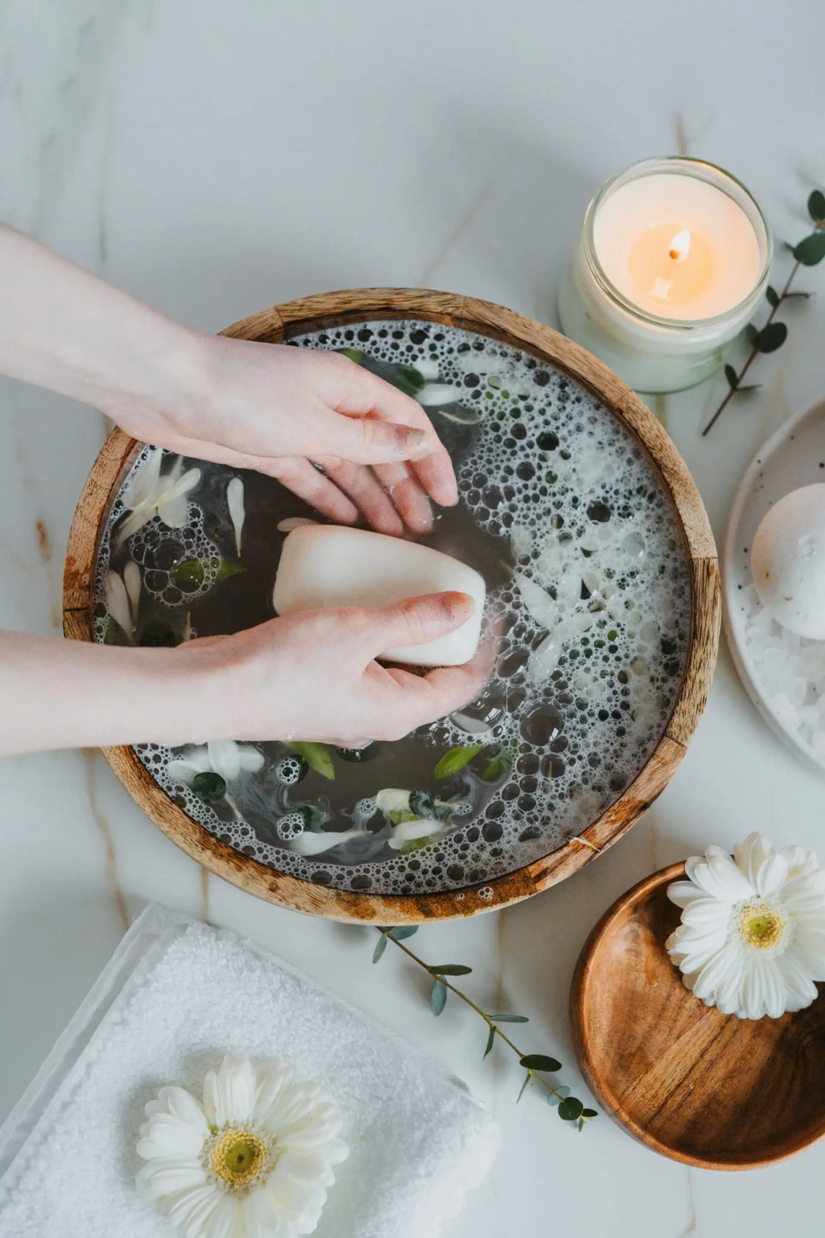 A pair of hands gently washing a bar of soap in a wooden bowl filled with soapy water and floating flower petals, with a lit candle and decorative items nearby.