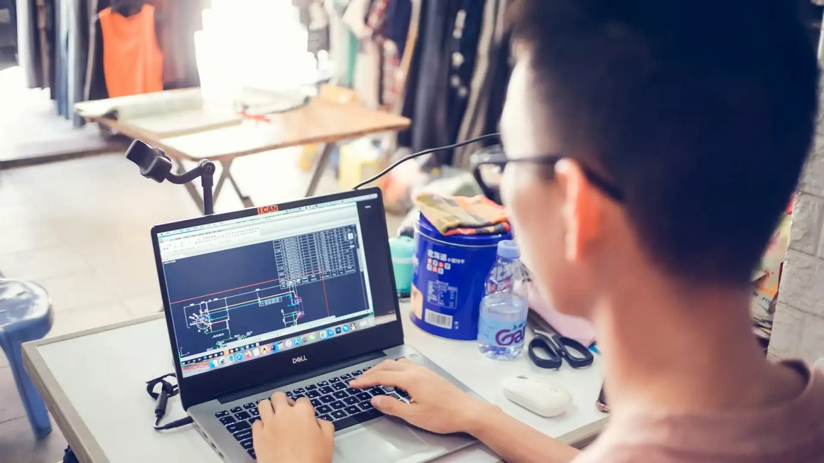 A person working on a laptop displaying technical drawings in a cluttered workspace.