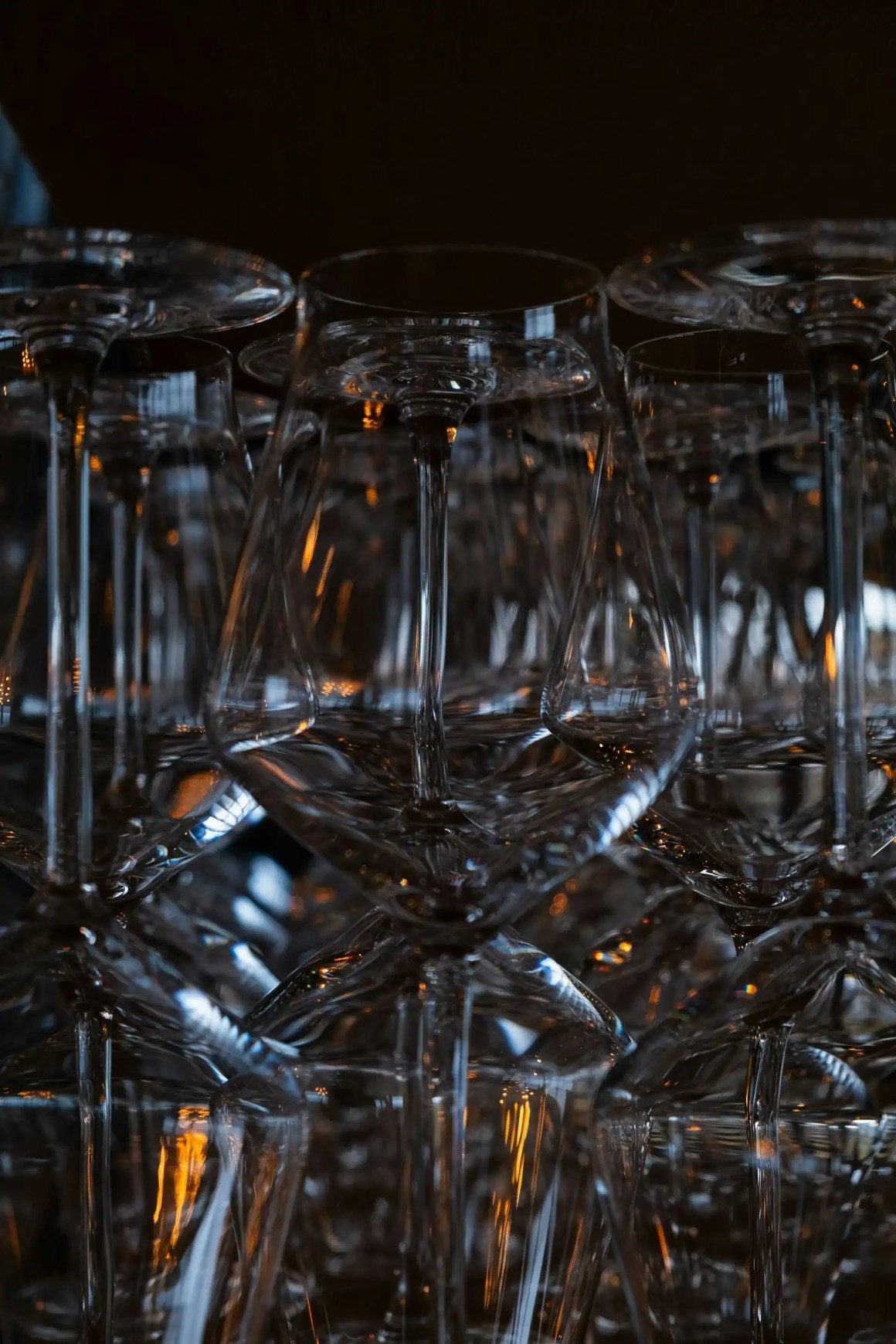 A close-up of stacked glassware, featuring wine glasses with shimmering reflections and warm highlights from the background.