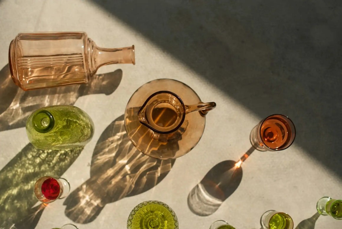 An overhead view of various colorful glassware, including green, pink, and amber bottles and glasses, casting intricate shadows on a light-colored surface.