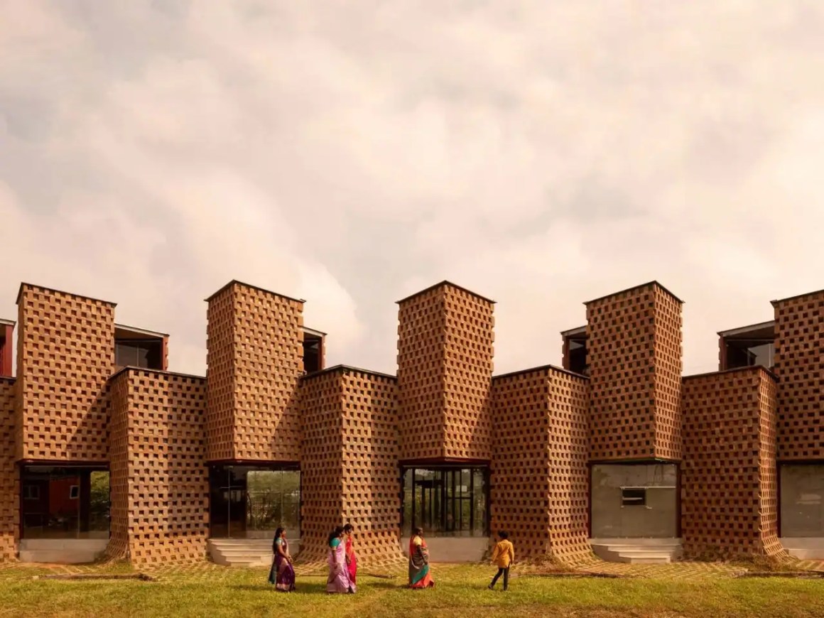 A modern architectural structure featuring a unique facade made of patterned clay bricks, with several figures walking in traditional attire in the foreground.