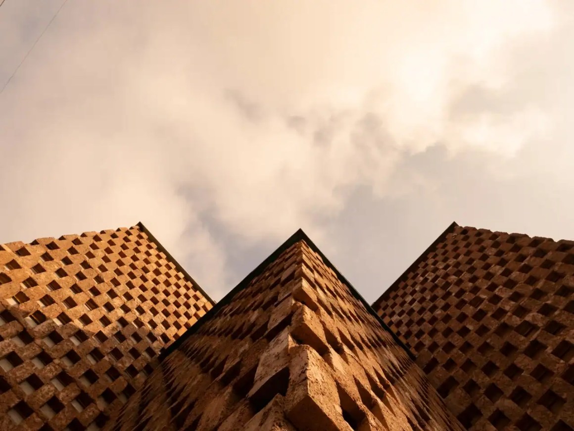 Looking up at two architectural structures with textured, patterned facades against a cloudy sky.