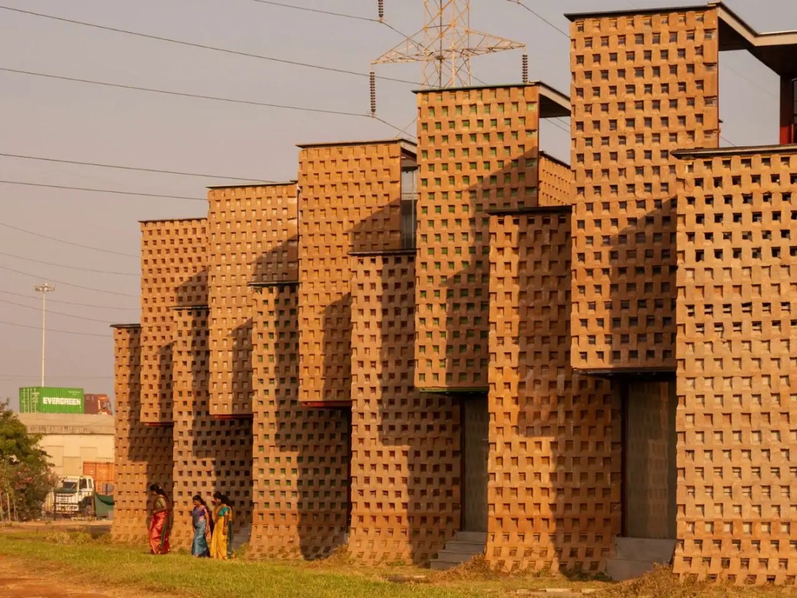 An artistic view of a modern building with a textured brick facade featuring square cutouts, surrounded by greenery and power lines in the background.