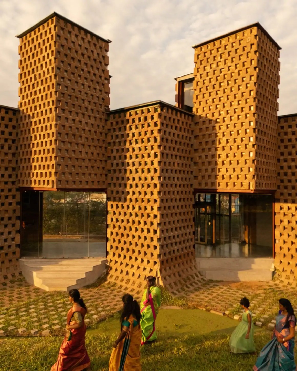 Exterior view of a modern building with textured brick walls featuring large opening windows, surrounded by green grass and paths, with several women in colorful traditional attire walking in the foreground.