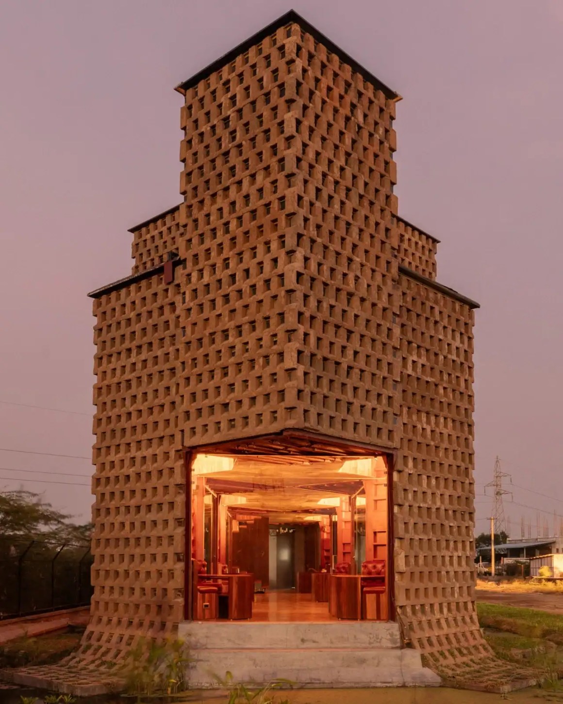 A distinctive brick building with a unique geometric design and glass entrance, illuminated from within, set against a twilight sky.