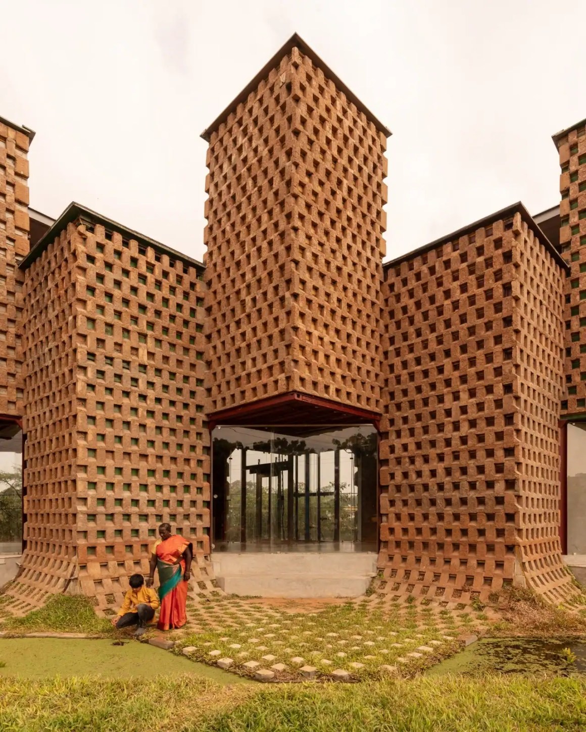 A modern building featuring a unique architecture of textured brick walls, with two people interacting in the foreground. One person is bending down while the other stands nearby, surrounded by green grass and pathways.