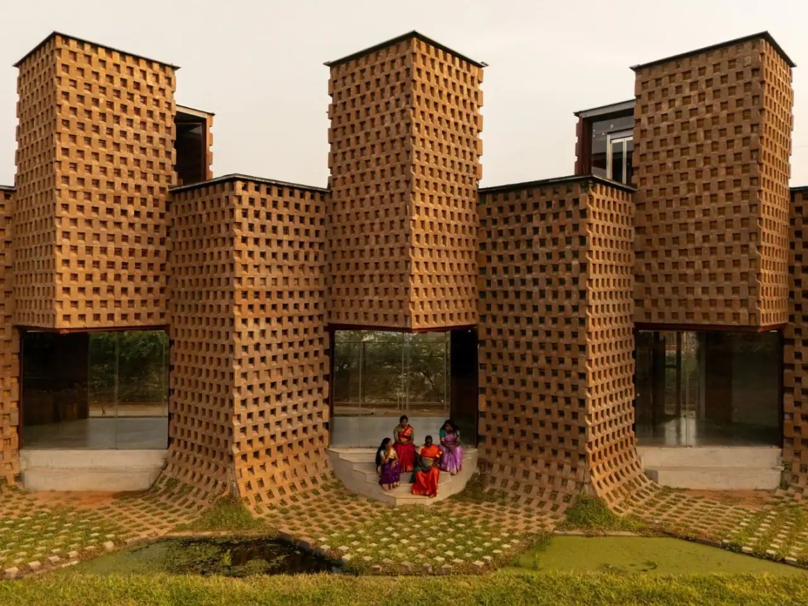 Three women in traditional attire sit outside a modern brick building with unique architectural features, surrounded by greenery.