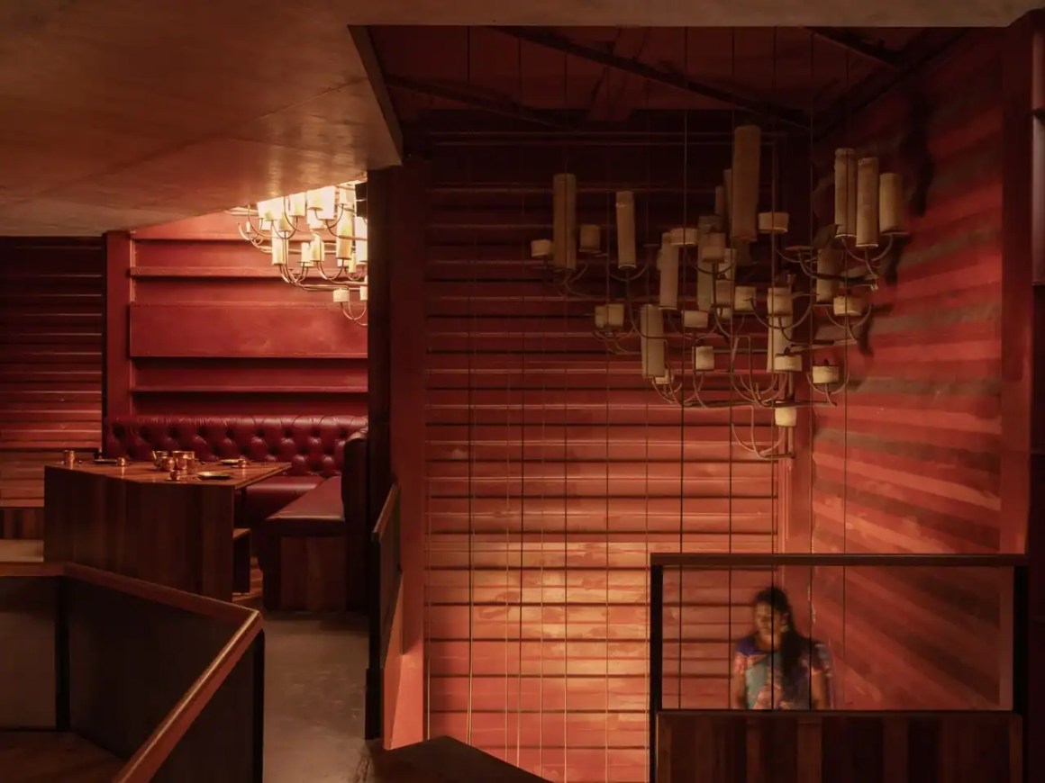 Interior view of a cozy restaurant featuring red wooden walls, a tufted seating area, and a modern chandelier, with a person standing in a stairwell.