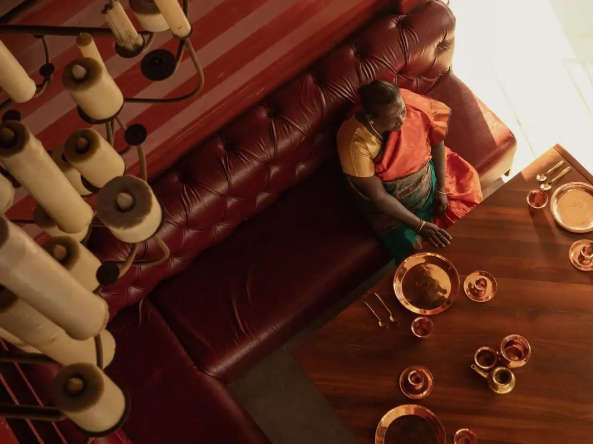 An overhead view of a woman seated at a table, surrounded by traditional Indian copper utensils and a decorative chandelier above.