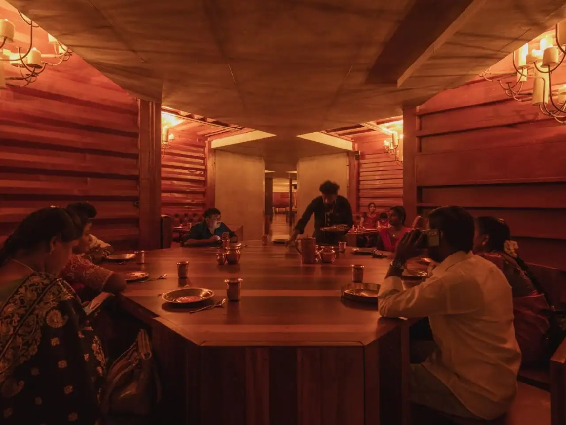 A dimly lit restaurant interior with a large wooden table surrounded by diners. The warm ambiance features decorative chandeliers and candlelight, creating a cozy atmosphere. Waitstaff is serving food, while some guests are engaged with their phones.