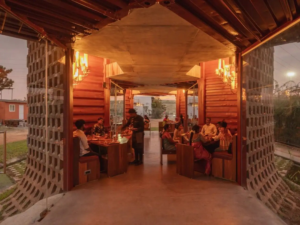 Interior view of a modern restaurant featuring warm lighting, wooden decor, and guests dining at tables. The design includes an open layout with large glass elements and a unique architectural structure.
